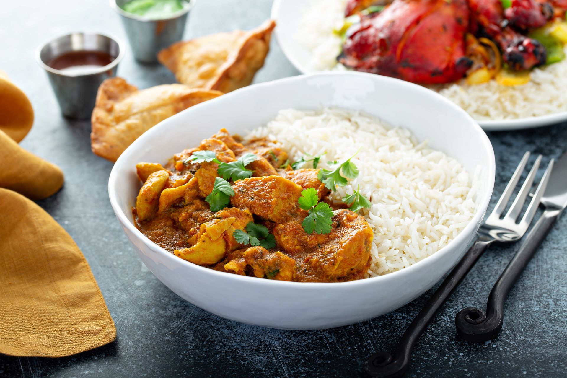 Chicken curry with white rice in a bowl, with naan, sauce, and pomegranate on a table.