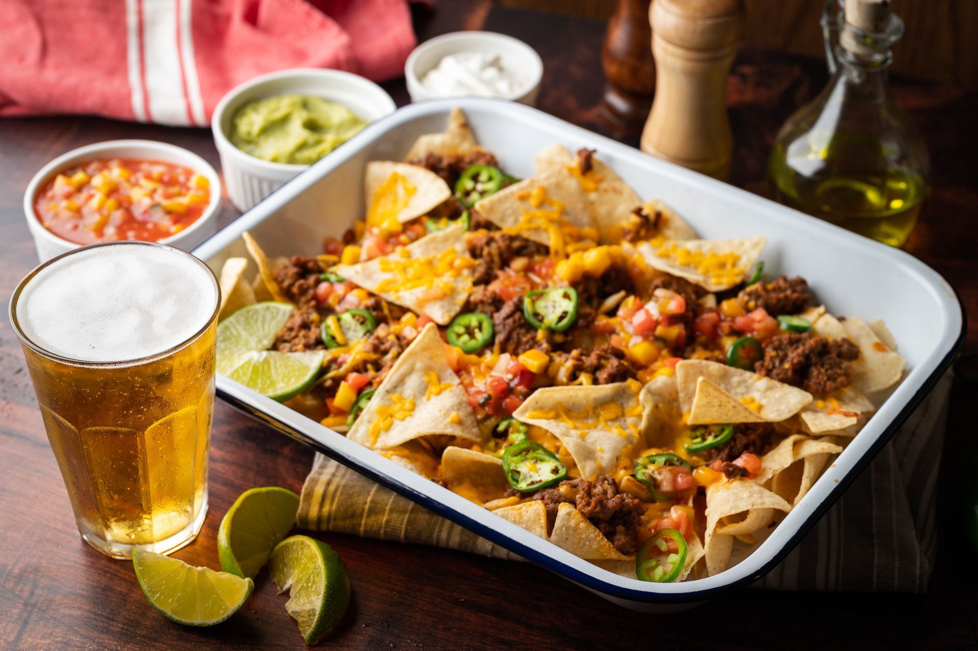Nachos in a white baking dish with beer, guacamole, salsa, and sour cream on a table