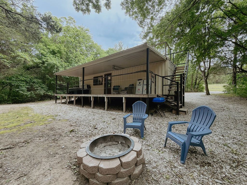Covered outdoor patio with a fire pit and blue chairs surrounded by trees and gravel.