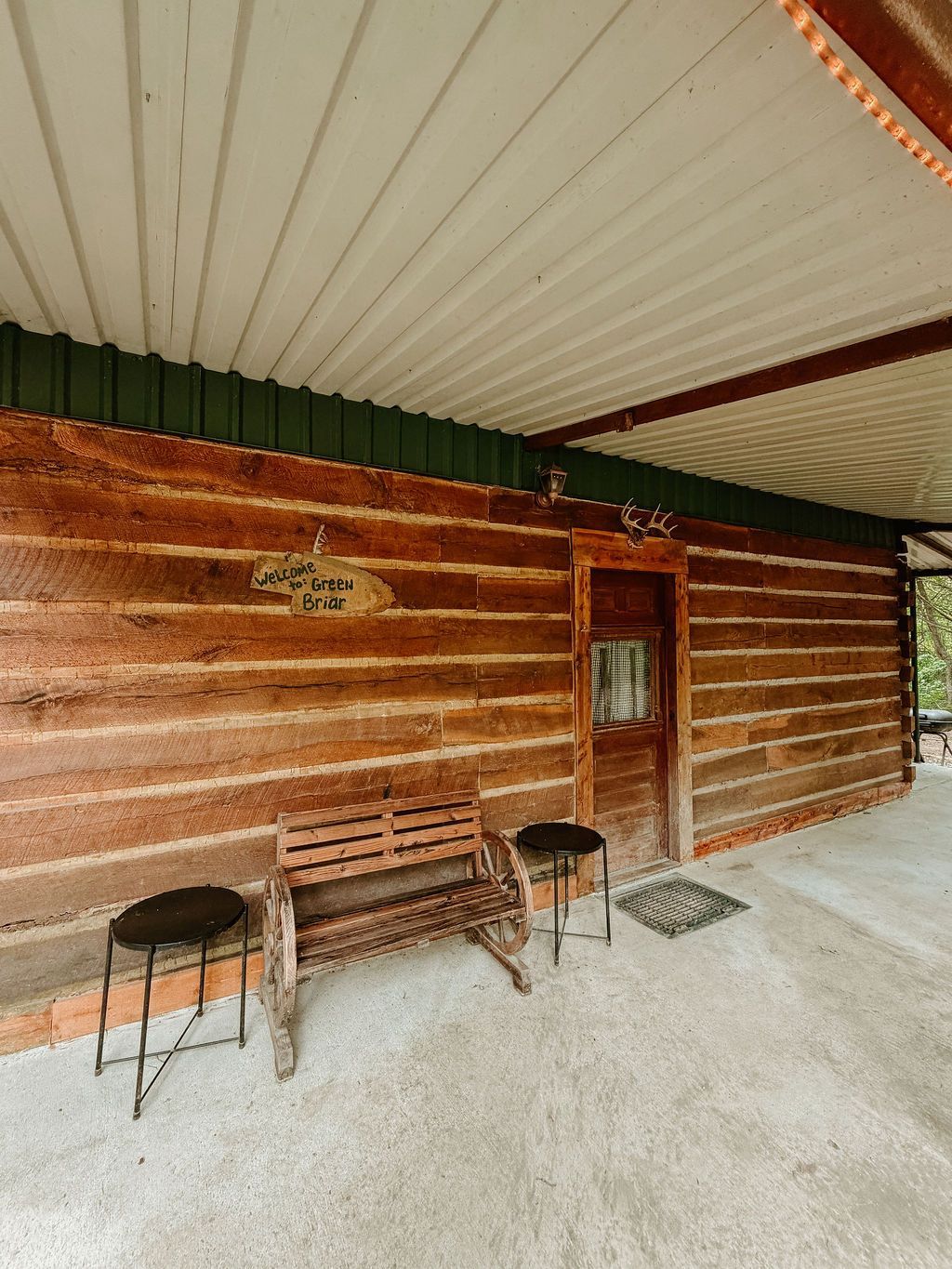 Rustic wooden porch with two stools, a bench, and a door under a corrugated metal roof.