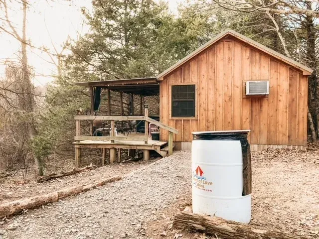 Rustic wooden cabin with a small porch and a white barrel in a rocky, wooded area