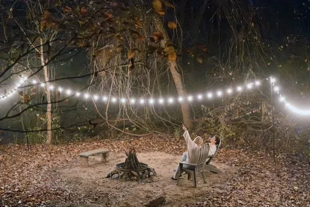 Person relaxing in a chair by a fire pit under string lights in a wooded backyard at night