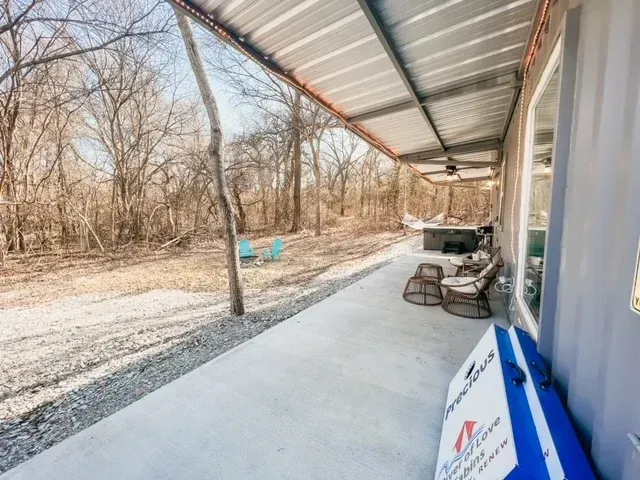 Covered concrete porch beside a wooded yard, with chairs and a blue cooler along the wall.