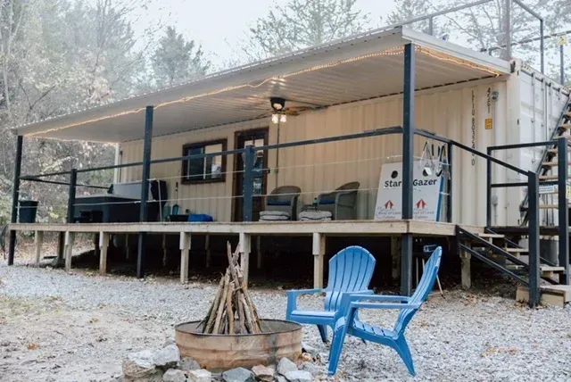 Rustic cabin porch with blue chairs, fire pit, and gravel yard in a wooded setting