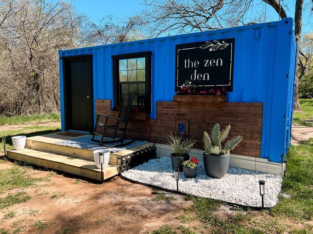Blue shipping container storefront with a porch, sign reading “the zen den,” and potted succulents