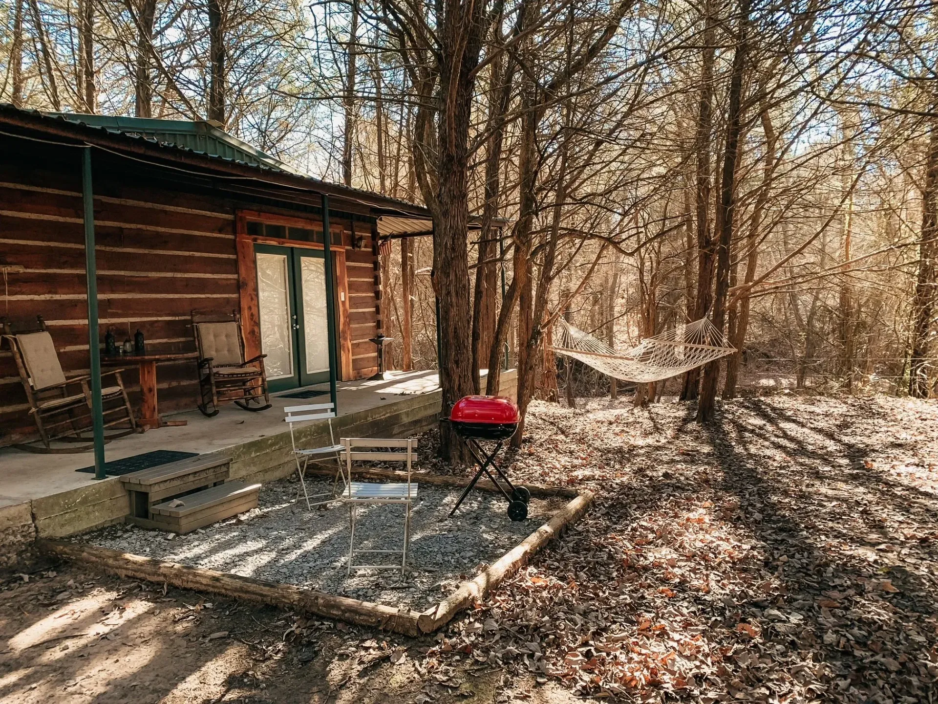 Rustic cabin patio with fire pit, chairs, hammock, and leaf-covered ground in a wooded area