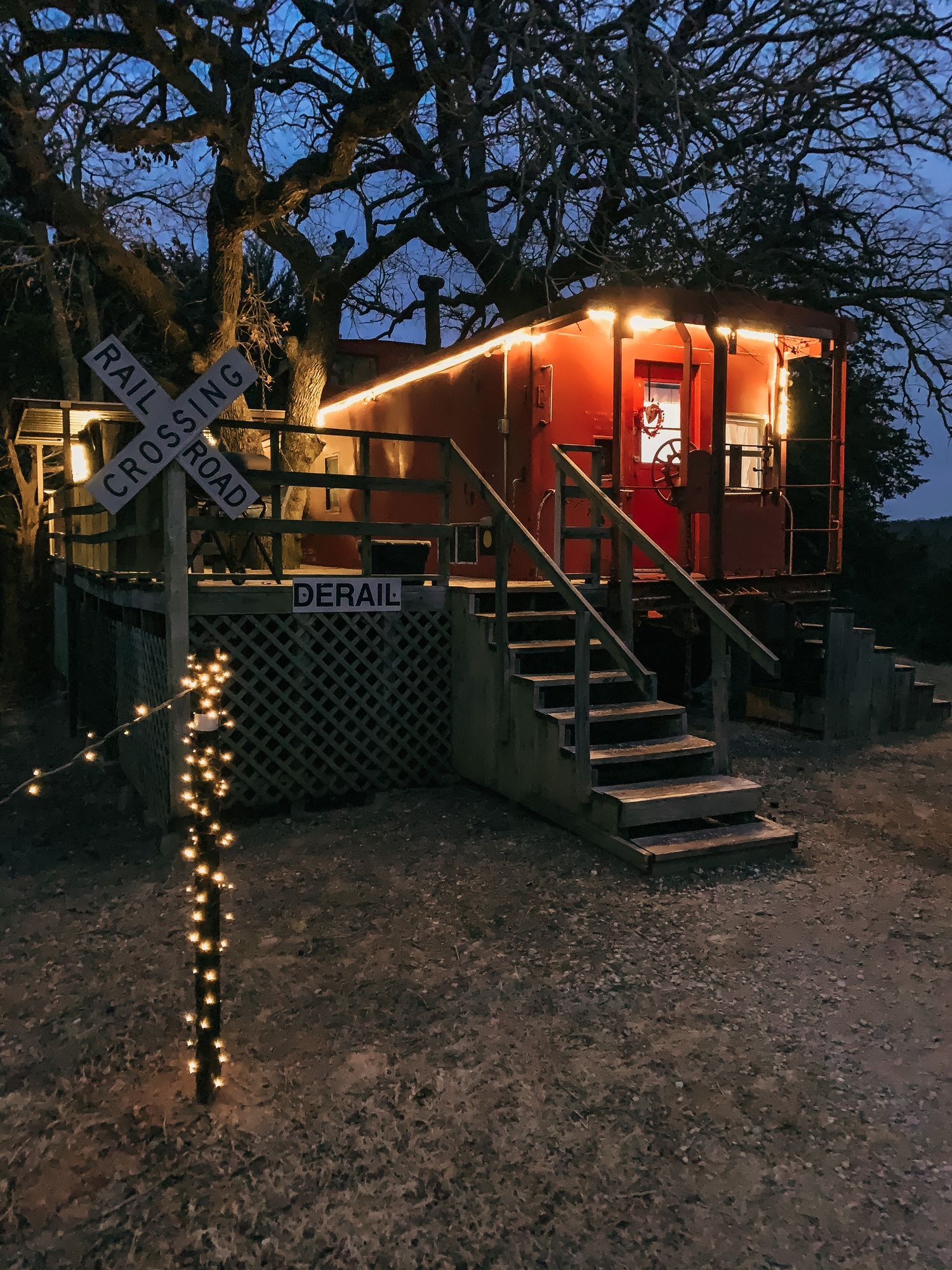 Red cabin lit at dusk beside a stairway and railroad sign, with string lights wrapped around a post.