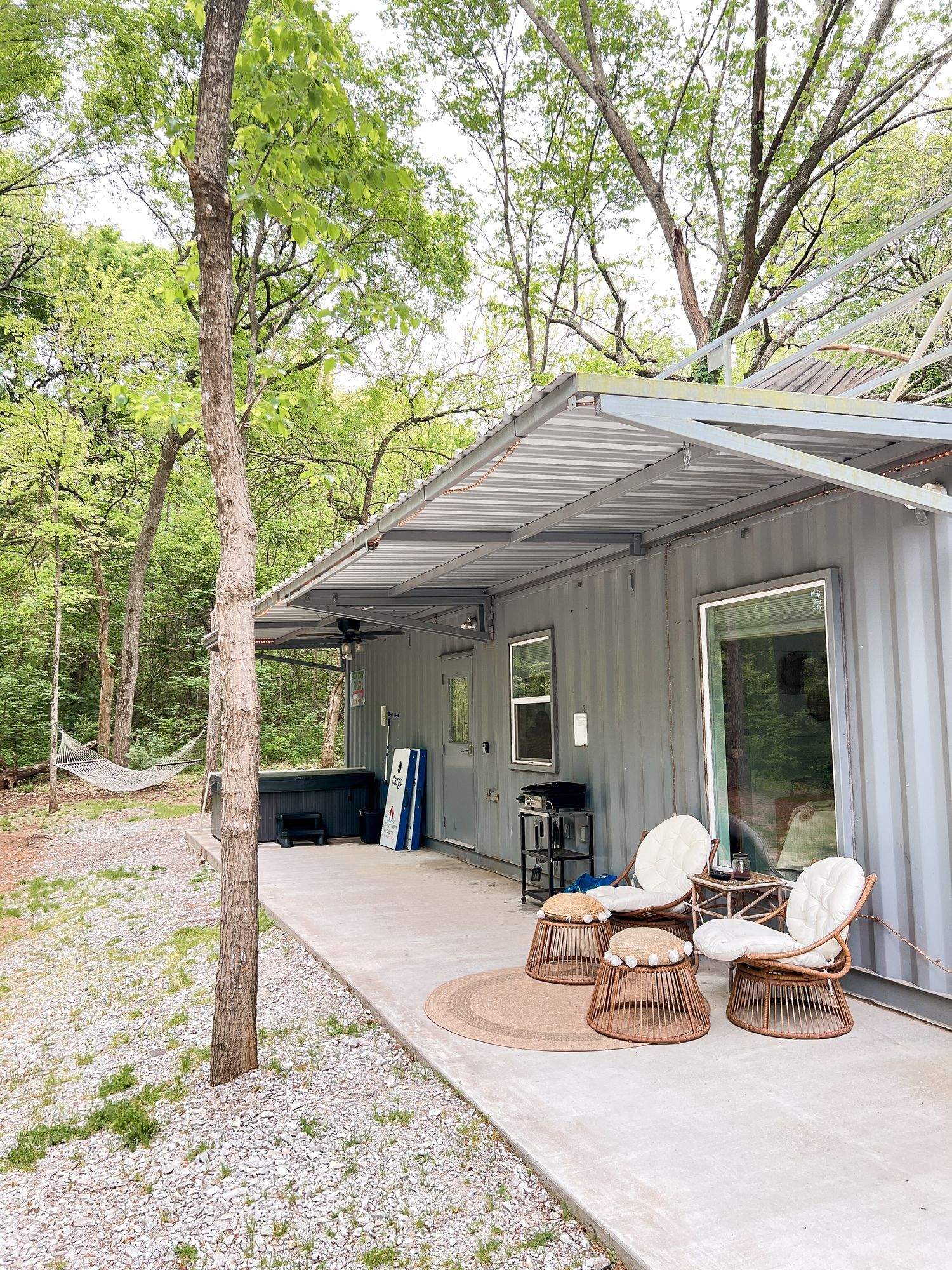 Outdoor cabin porch with metal awning, chairs, table, and gravel path beside trees