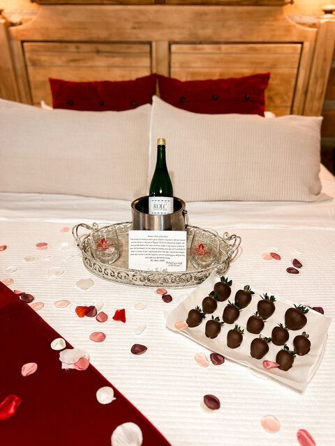 Champagne bottle with chocolates and rose petals arranged on a bed with white linens