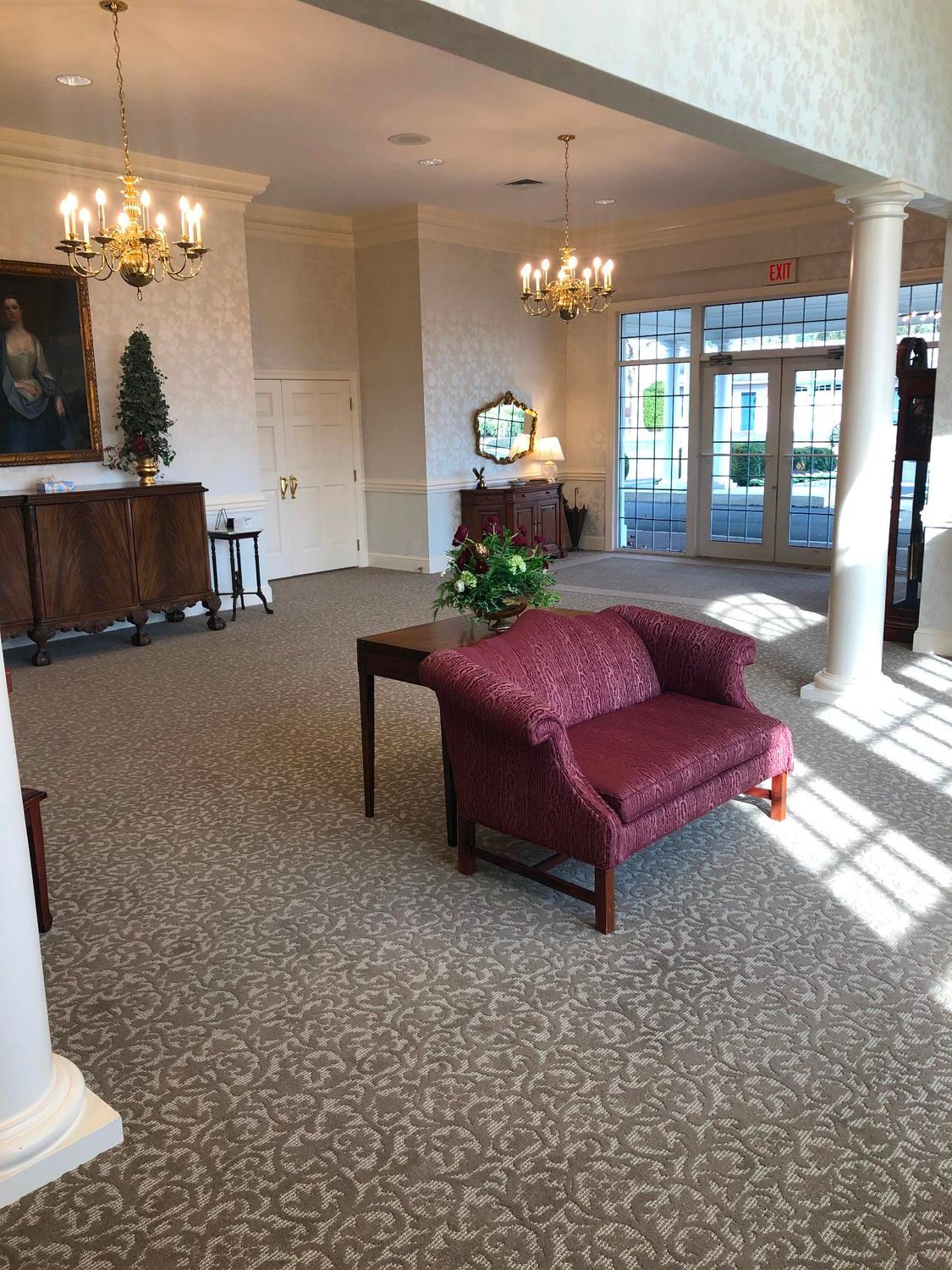 Empty hotel lobby with patterned carpet, red chaise lounge, chandelier, and glass entrance doors