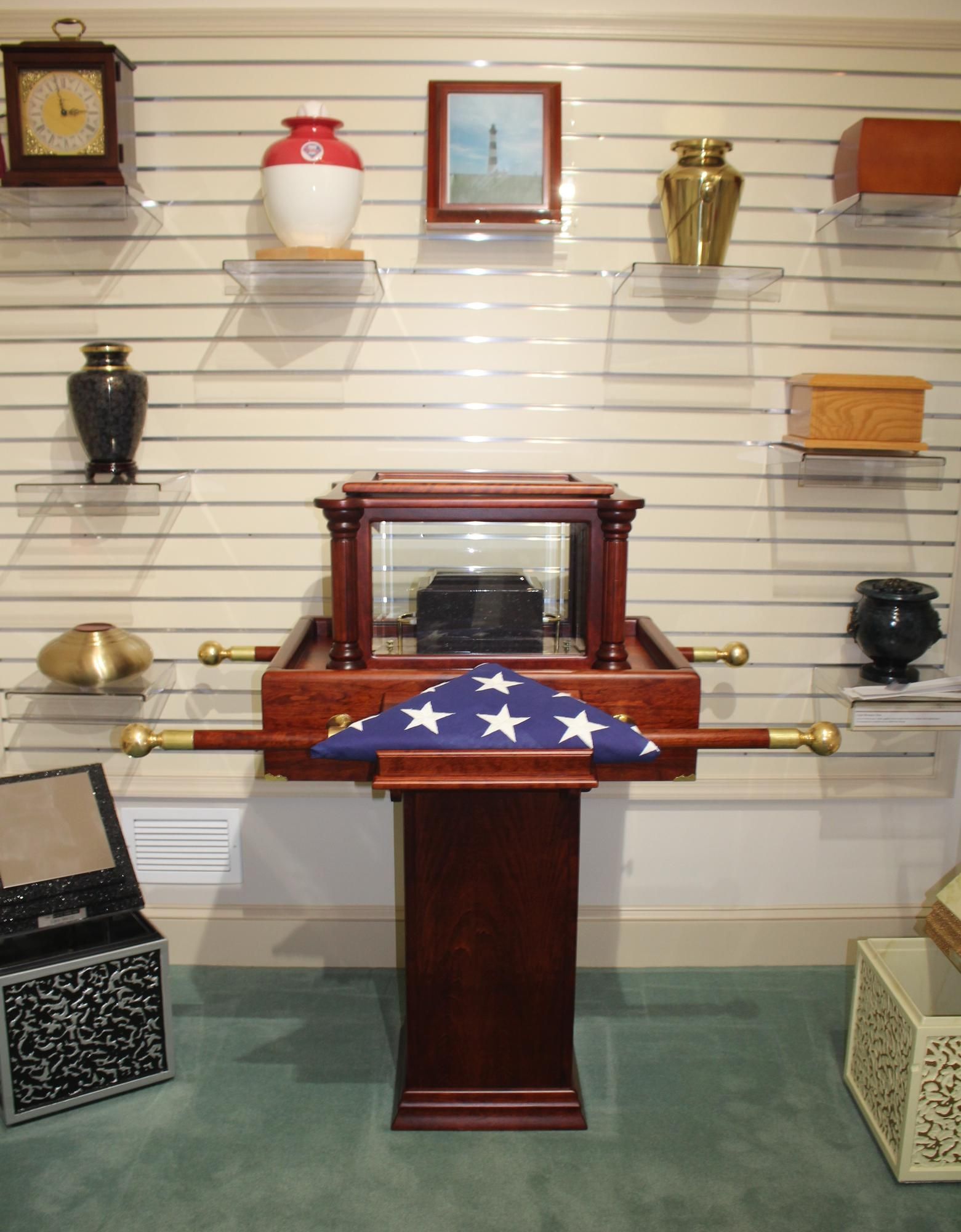 Coffin draped with a U.S. flag on a pedestal in a funeral home showroom, surrounded by urns and caskets