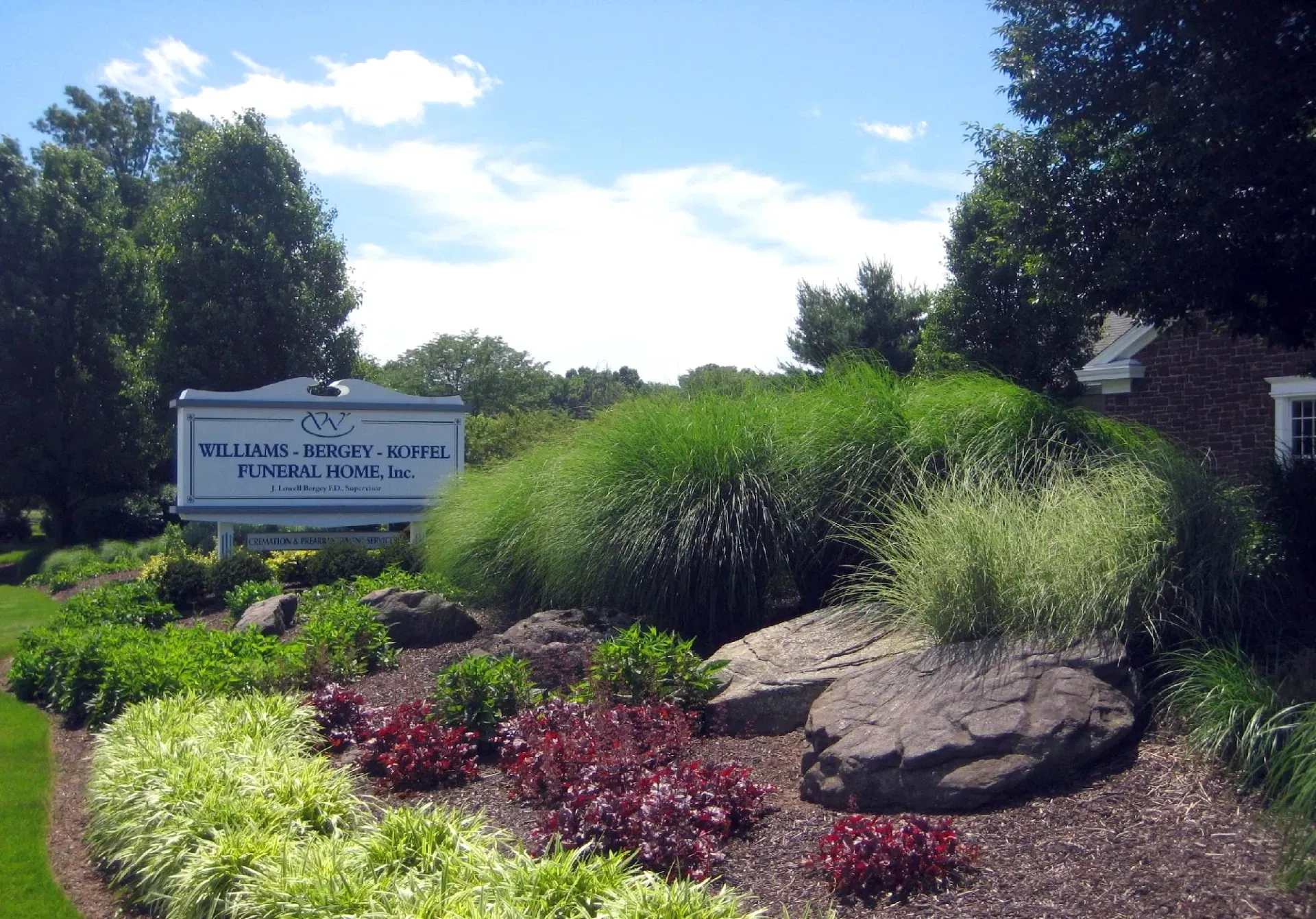 School sign beside landscaped garden with rocks, flowers, and tall green shrubs under a sunny sky