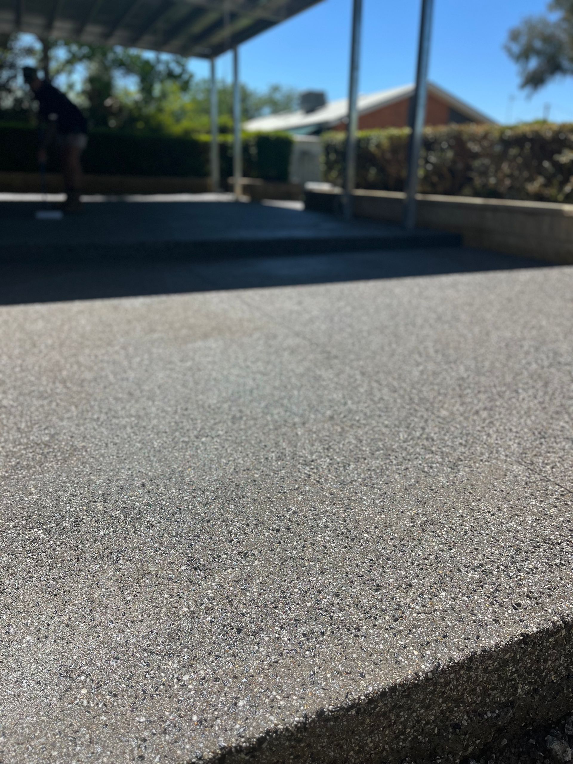 Sunlit outdoor court with a textured gray surface, covered area, and houses in the background — Complete Concreting Riverina In Ashmont, NSW