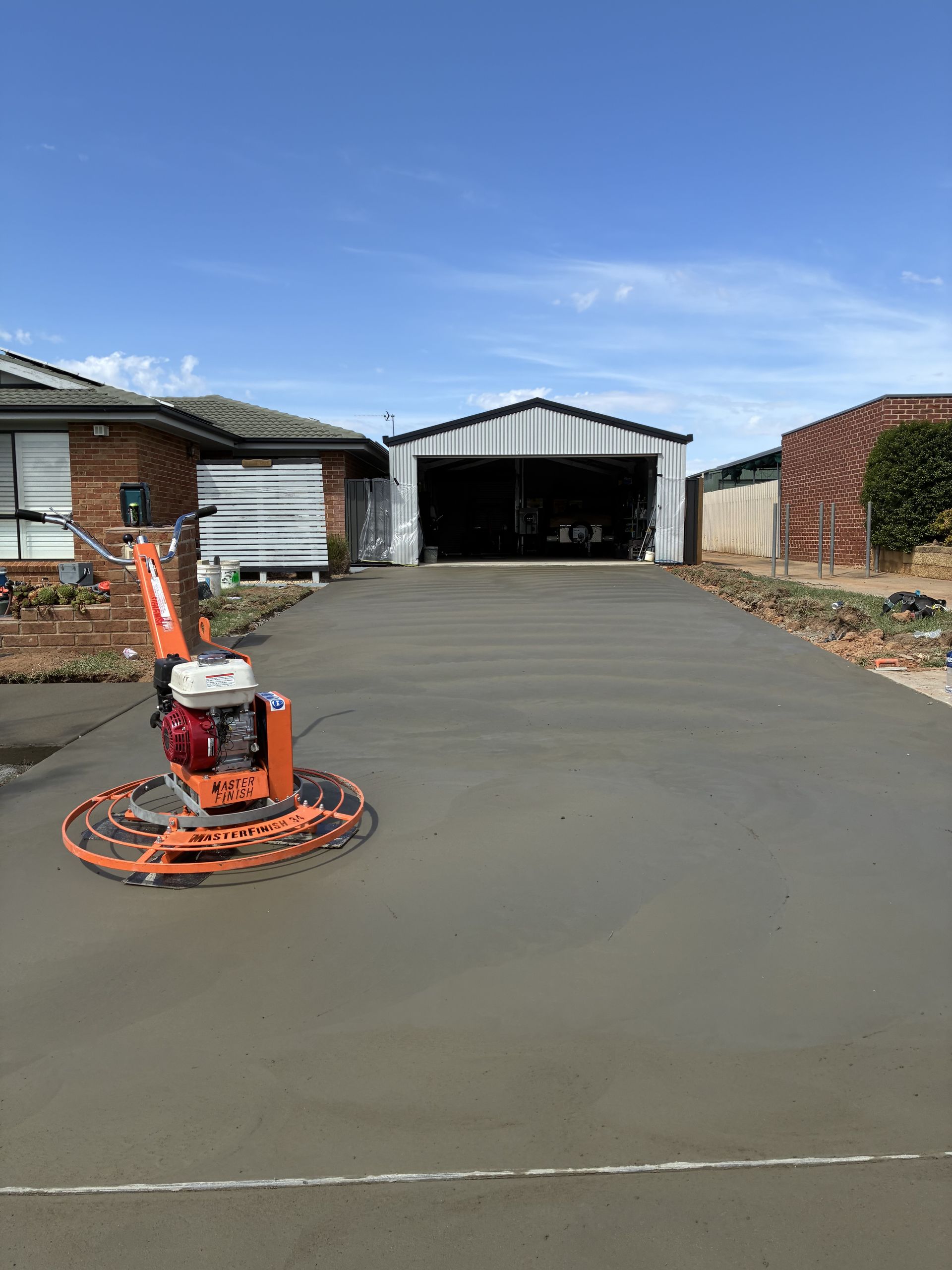 Orange concrete float on fresh driveway in front of suburban garages under a clear blue sky — Complete Concreting Riverina In Ashmont, NSW