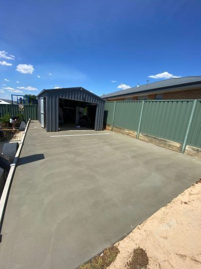 Freshly poured concrete driveway beside a green fence and gray shed under a blue sky — Complete Concreting Riverina In Ashmont, NSW