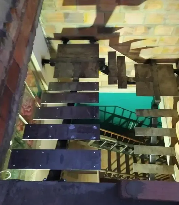 Escalera interior estrecha con escalones oscuros y barandilla metálica junto a una pared de ladrillo y una puerta turquesa