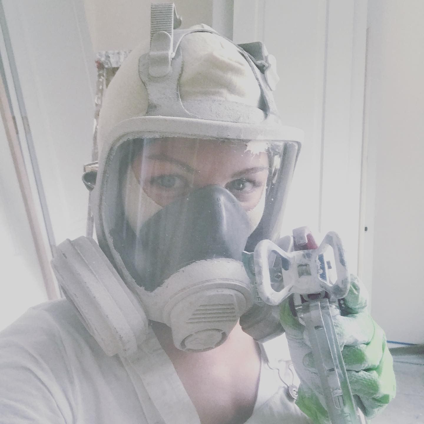 A person wearing a full-face respirator and paint-covered gloves holds a paint sprayer in an indoor setting.