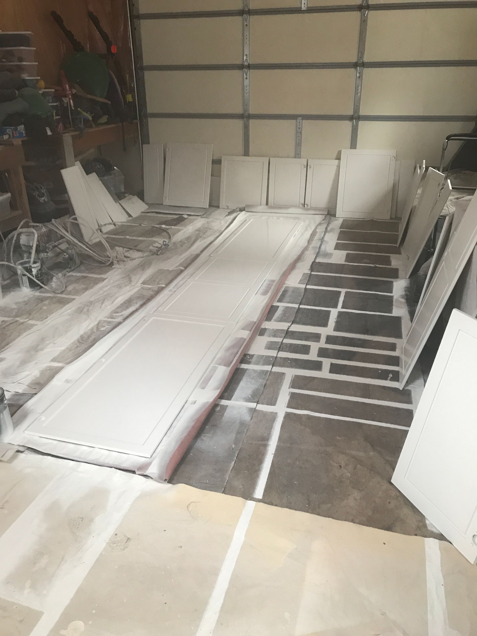 Several white cabinet doors are laid out on plastic sheeting on a garage floor to be painted.