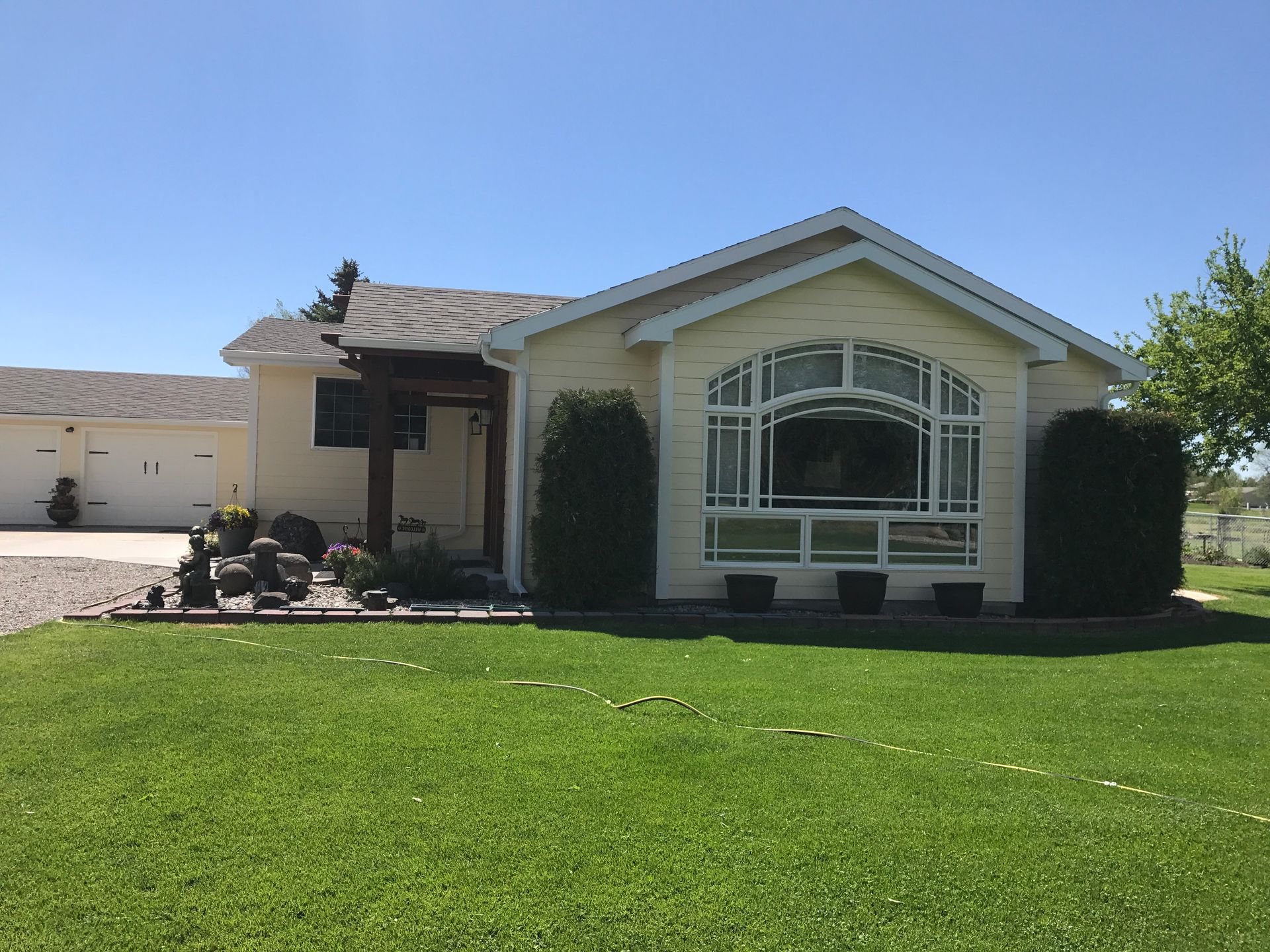 A single-story, light yellow house with a prominent arched window and a covered entryway, set behind a lush green lawn.
