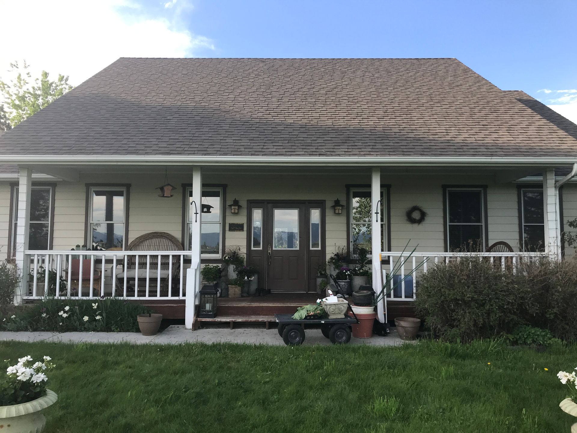 A house with a beige exterior, a brown shingled roof, a front porch with a white railing, and a central brown door.