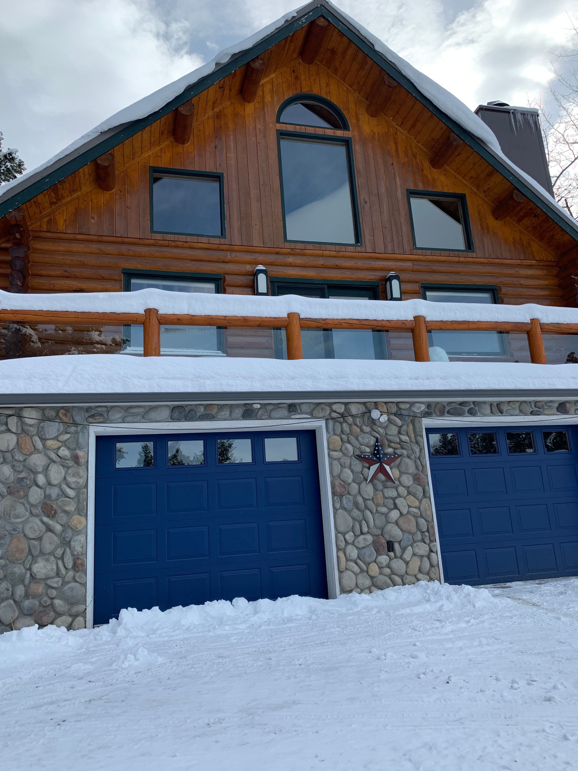 A log cabin with a blue two-car garage base and a snowy balcony, set against a winter landscape.