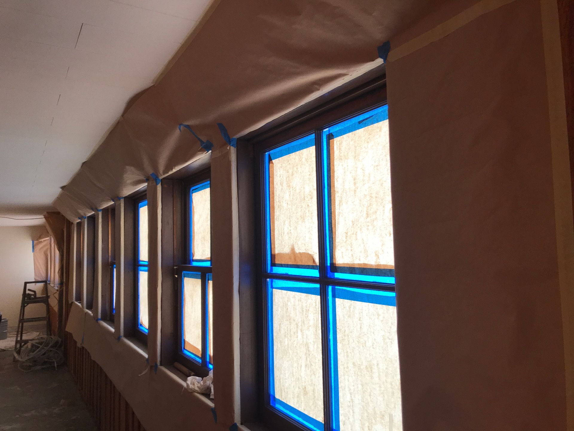 A row of windows with blue painter’s tape and brown masking paper covering the trim, prepared for painting.
