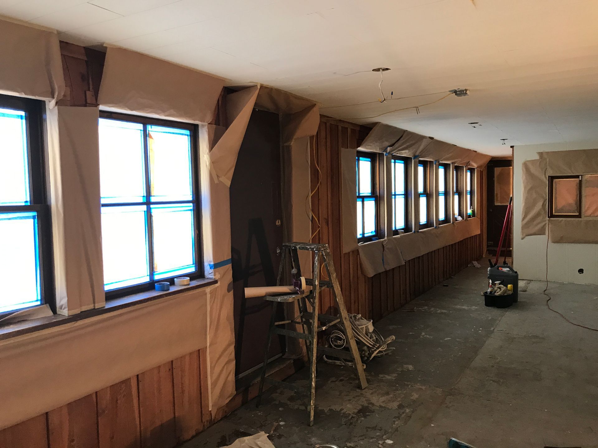 An interior room under renovation with walls and windows masked with brown paper and a step ladder in the center.