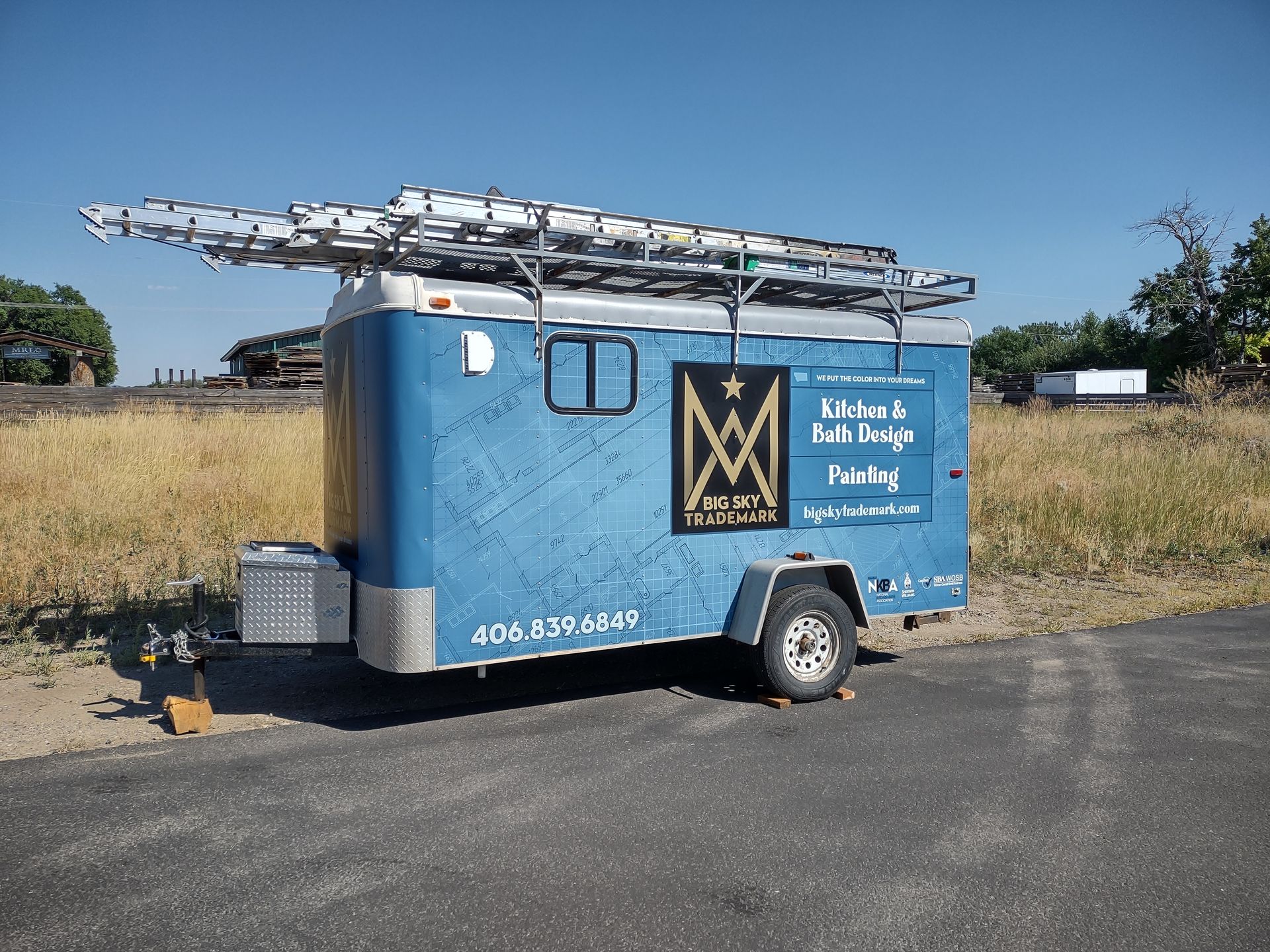 A blue utility trailer parked on asphalt, featuring a business logo, a ladder rack on top, and a clear blue sky background.
