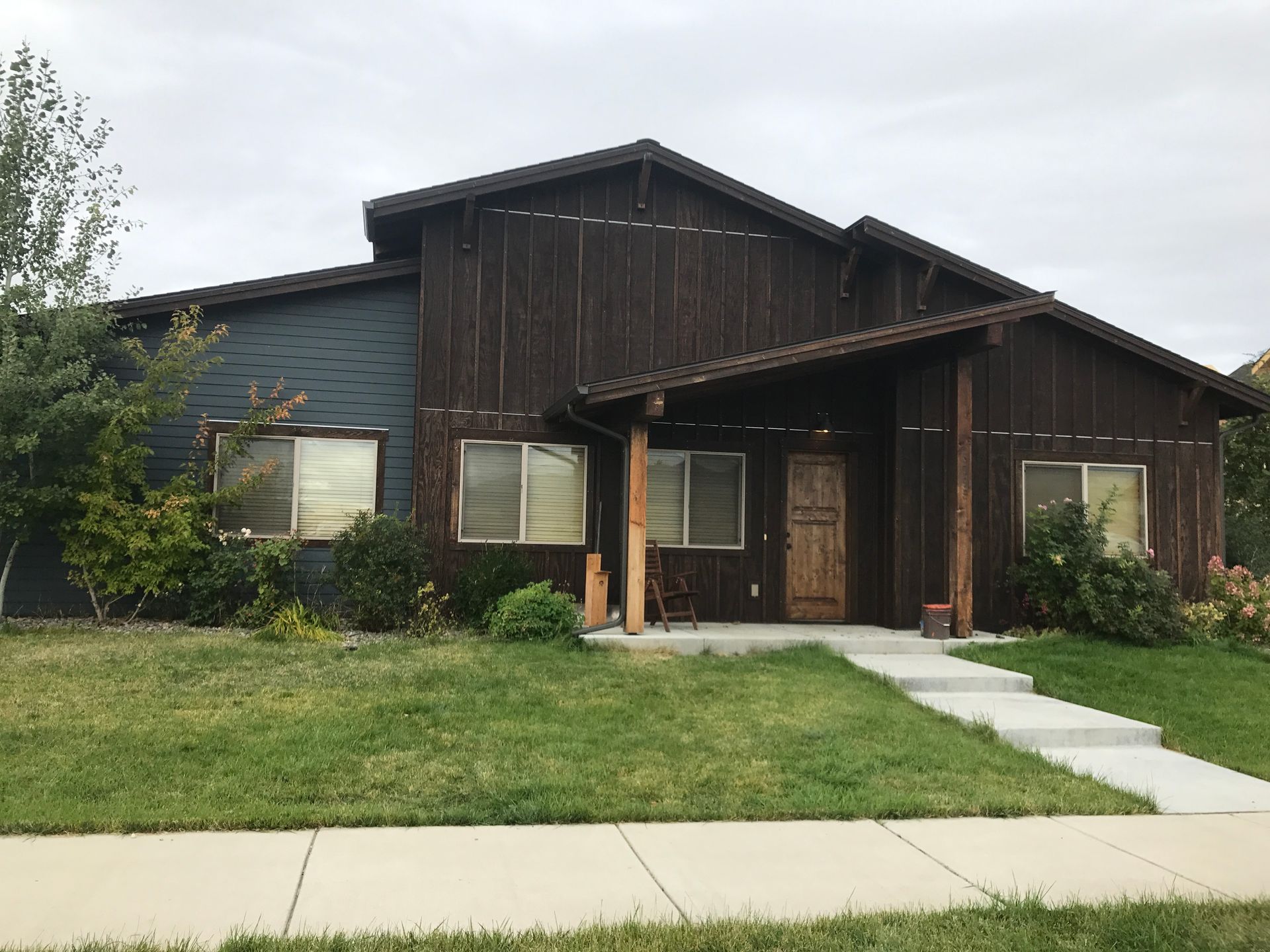 A dark wood-paneled single-story house with a prominent front porch, concrete walkway, and lawn under a cloudy sky.
