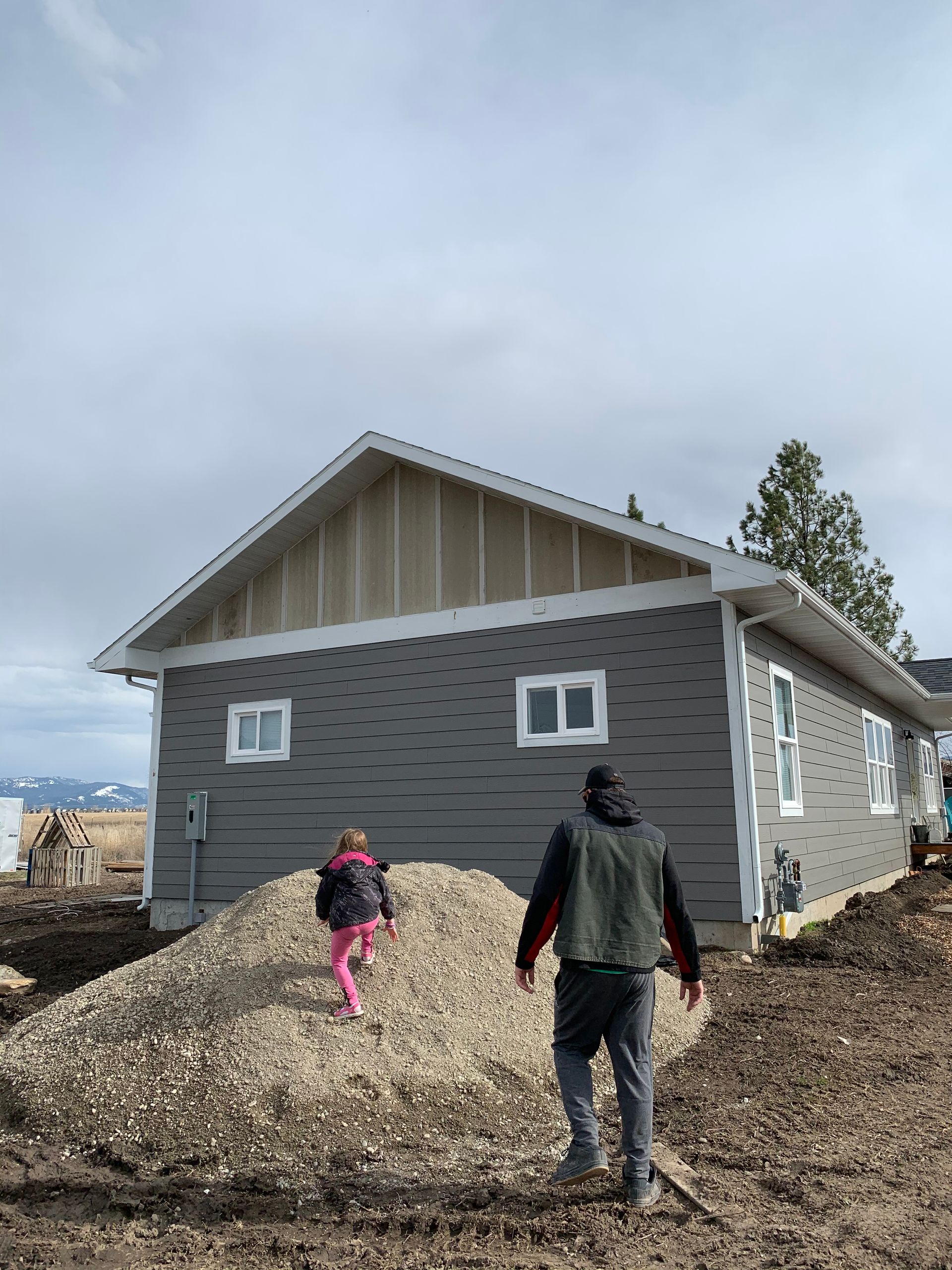 A person and a child walk toward a house under construction with grey siding and a gravel pile in front.