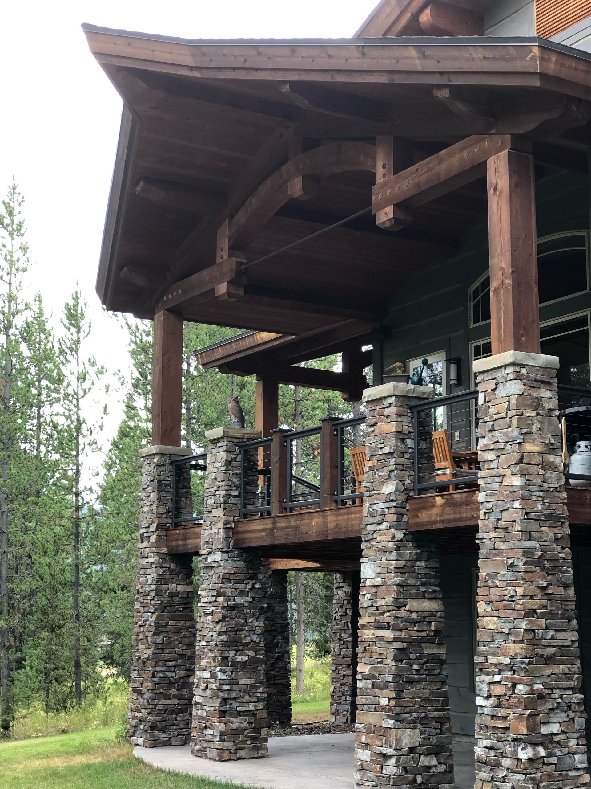 A rustic cabin deck features stone pillars supporting a wooden overhang, set against a background of pine trees.