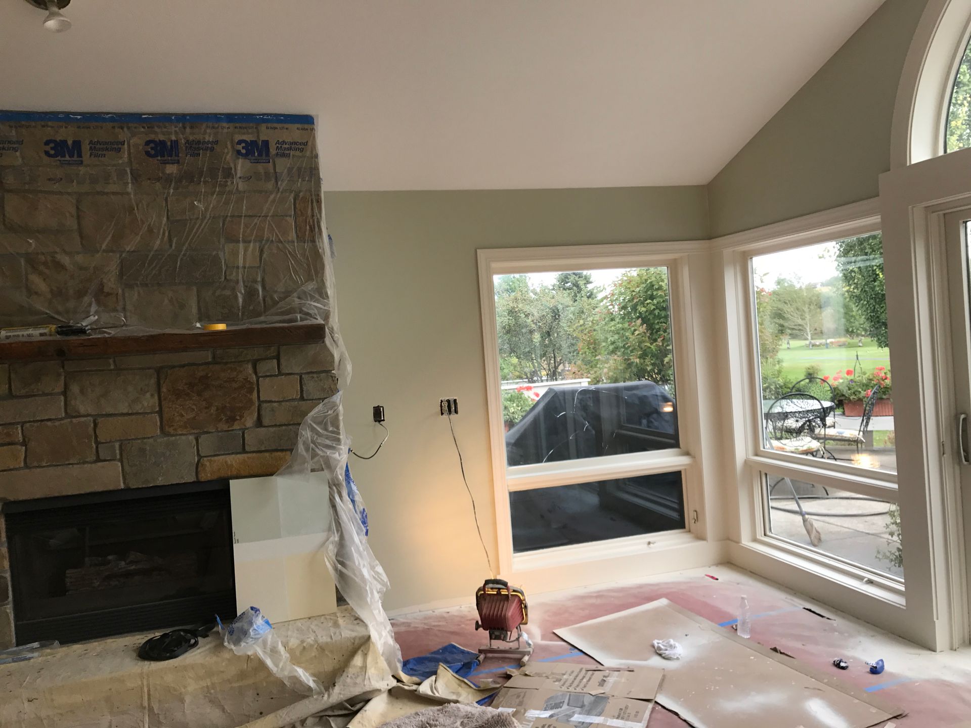 A room under renovation with a stone fireplace covered in plastic, light green walls, and large windows.