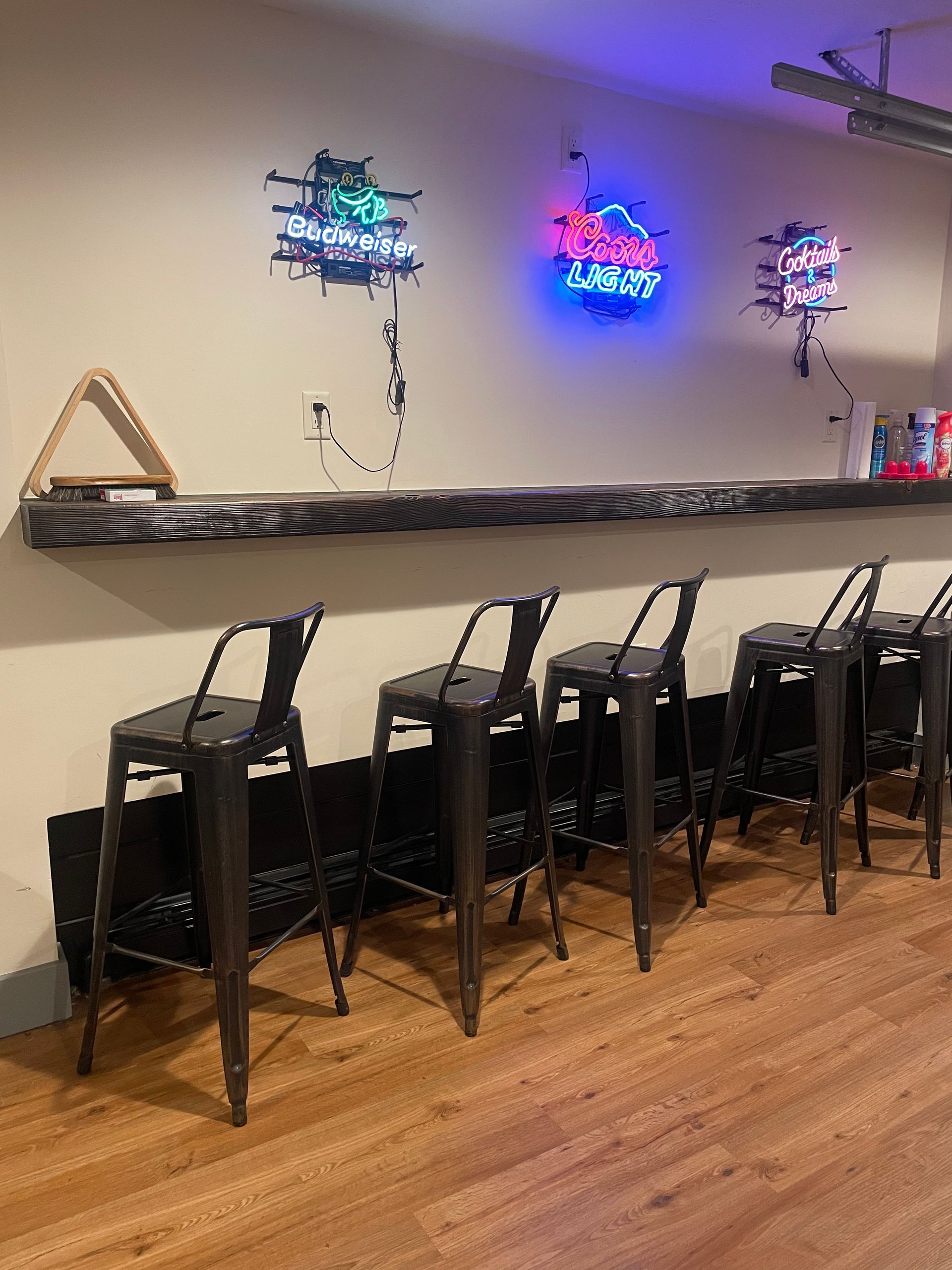 A row of five black metal bar stools lined up against a long, wooden bar top with neon signs mounted on the wall above.