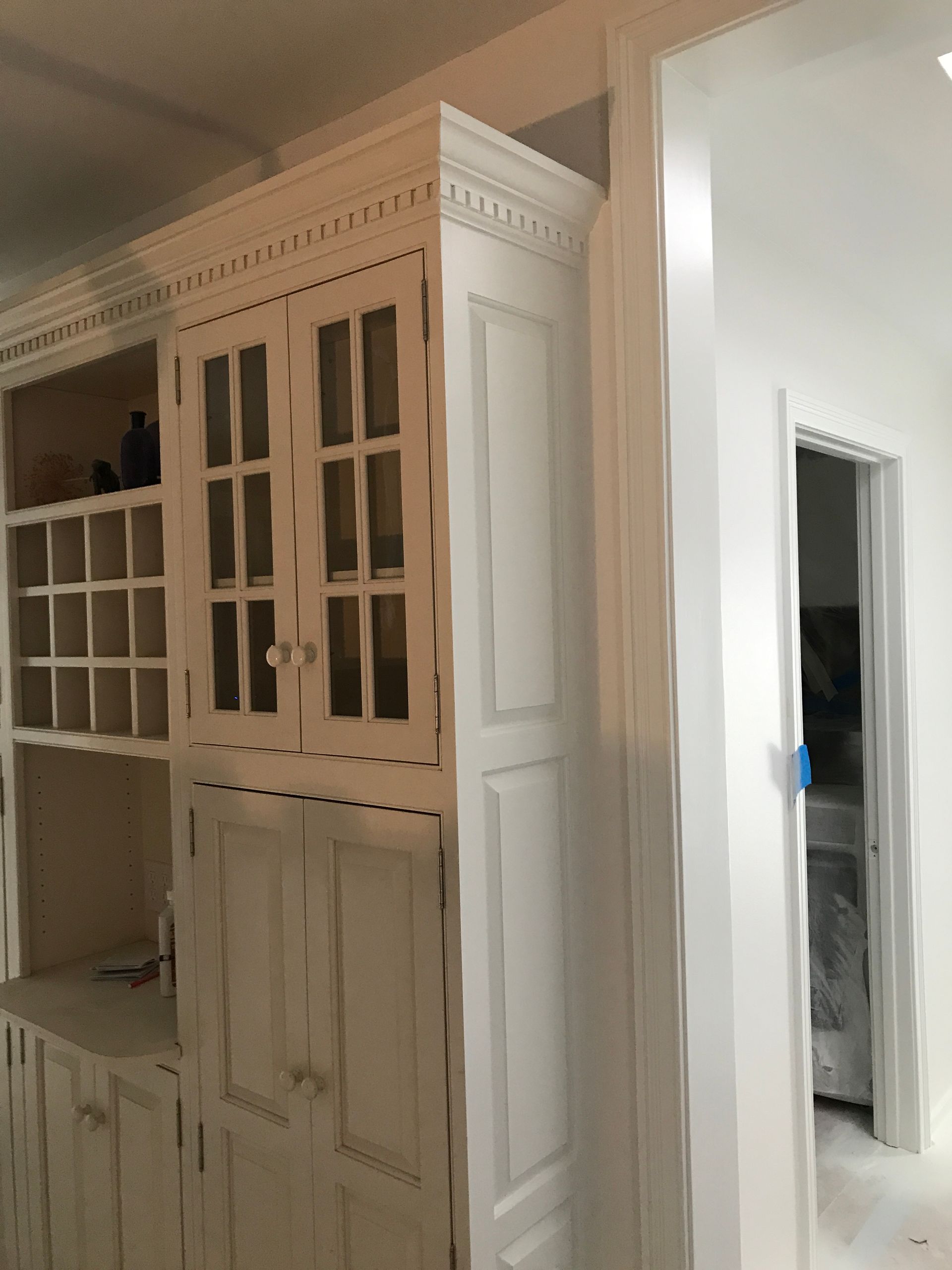 A white cabinet with glass upper doors and wine storage next to a doorway in a brightly lit, neutrally decorated room.