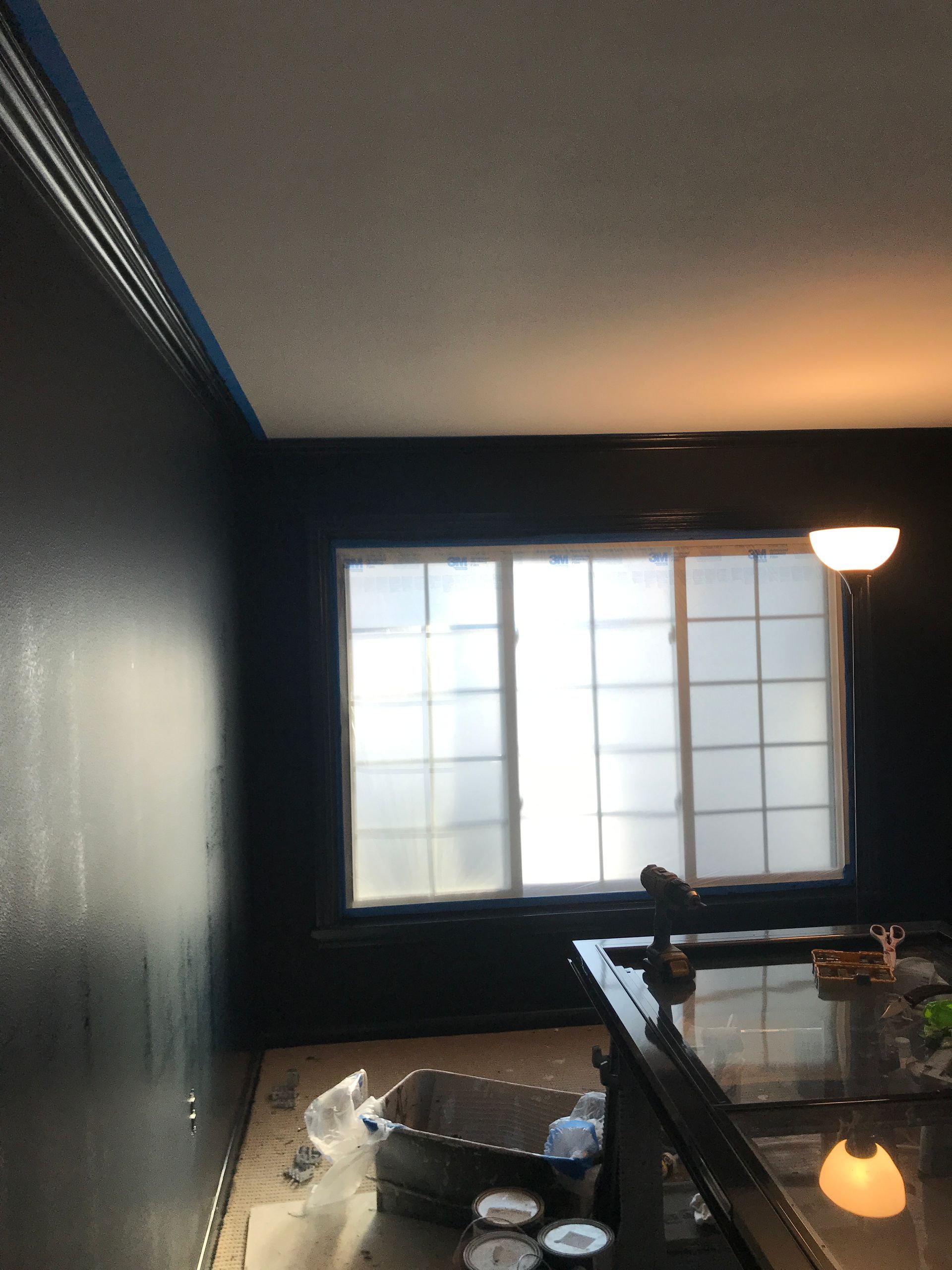 A dark-walled room undergoing renovation, featuring blue painter's tape along the ceiling and a large window.