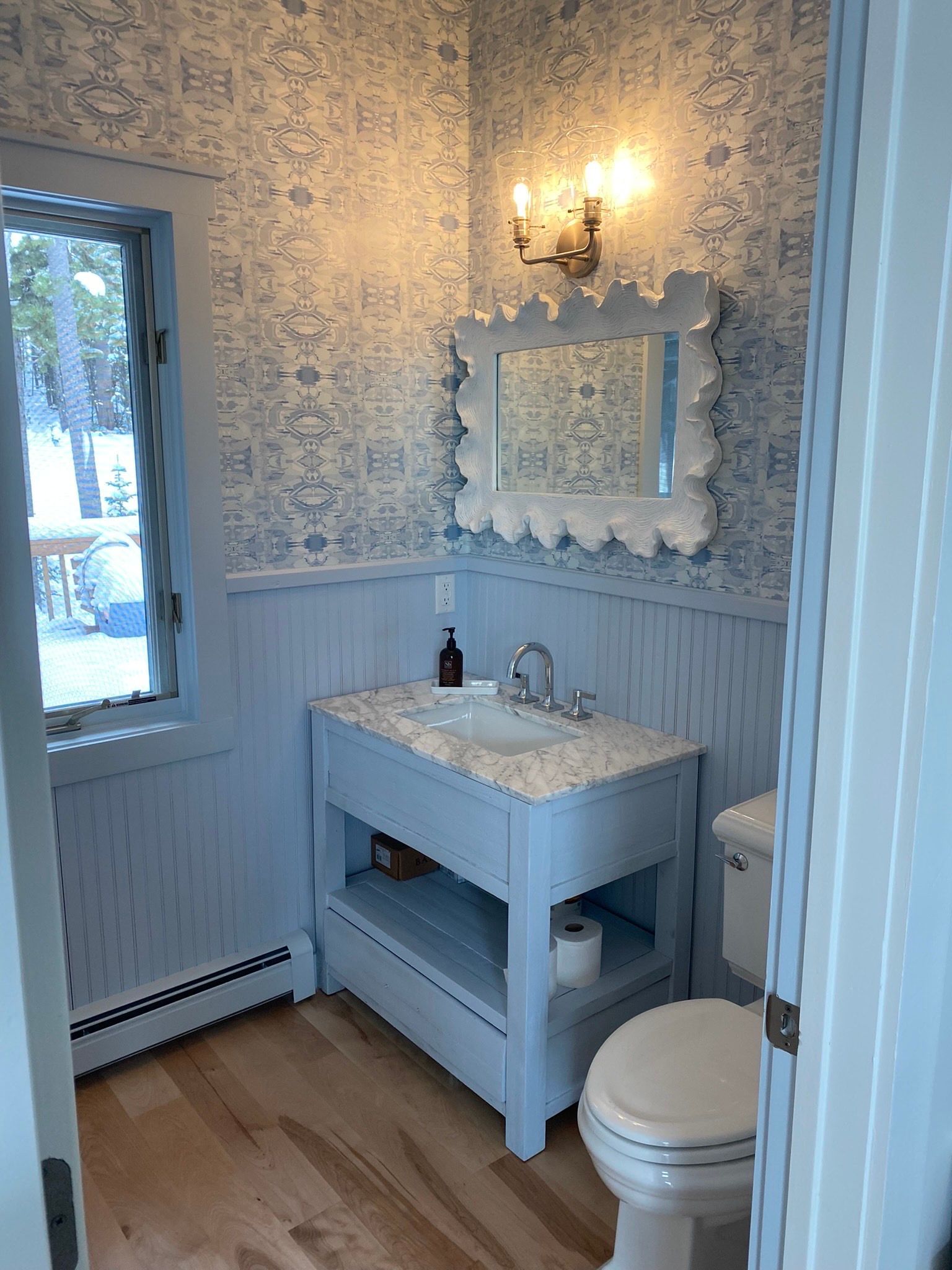 A small bathroom with light blue beadboard wainscoting, patterned floral wallpaper, a vanity, and a white framed mirror.