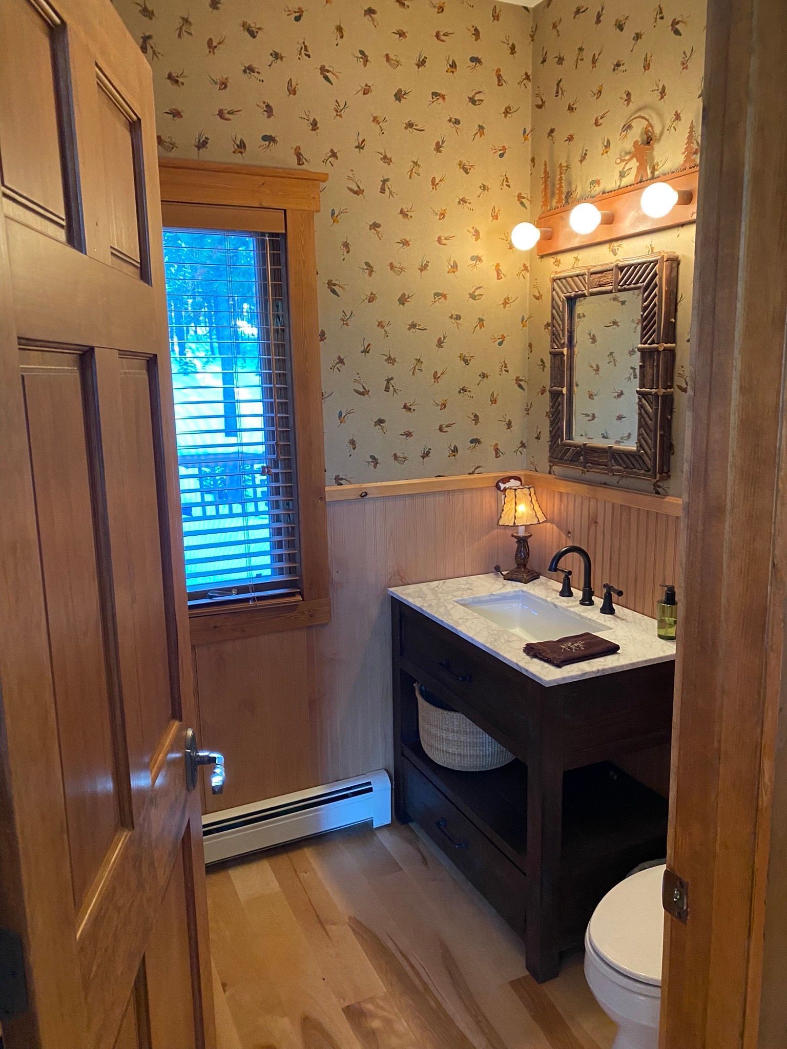 A small rustic bathroom with floral wallpaper, wood paneling, a wooden vanity with a white sink, and a framed mirror.