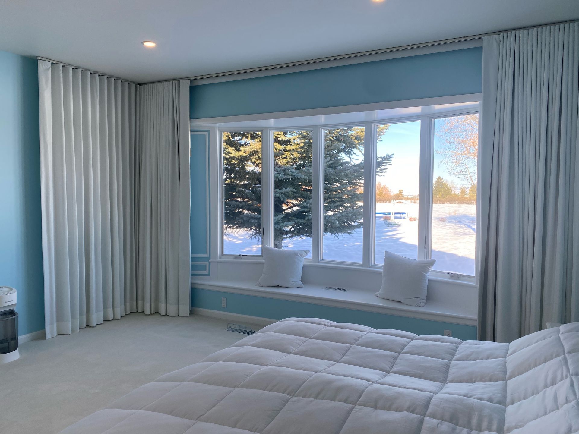A bright bedroom features a window seat looking out onto a snowy yard, framed by floor-to-ceiling light gray curtains.