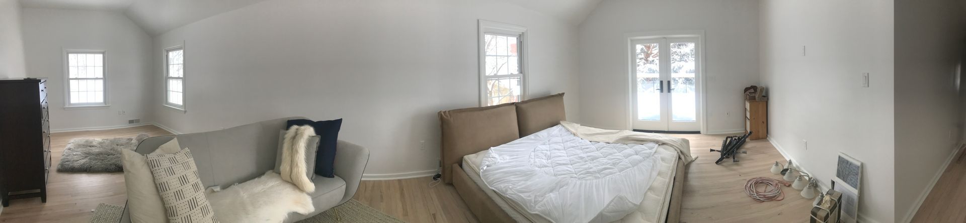 A panoramic view of a bedroom with light wood floors, a sofa, a bed with tan headboards, and French doors.