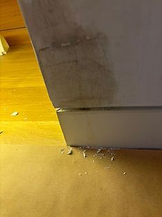 A close-up of a white painted baseboard with dark scuff marks, a horizontal crack, and white debris on the wooden floor.