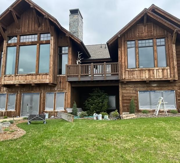 A large, two-story wooden house with dark timber siding, a stone chimney, and a central balcony overlooking a lawn.