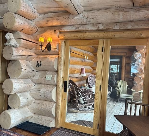A rustic log cabin wall with a double glass door opening onto a deck with chairs and a small decorative dog figurine.