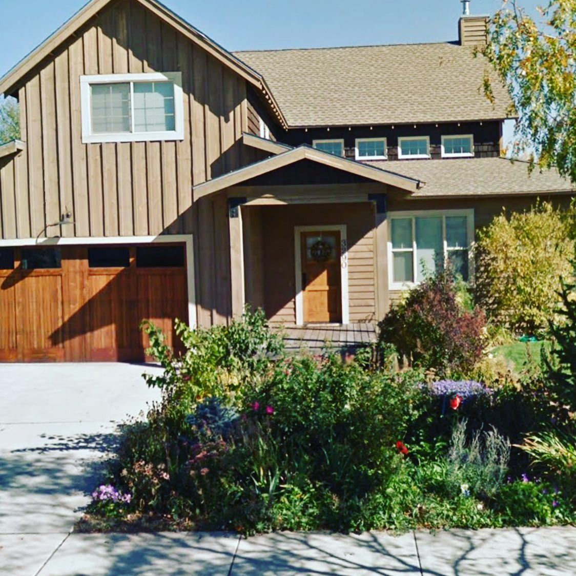 A two-story brown house with vertical siding, a wooden garage, and a landscaped front garden on a sunny day.