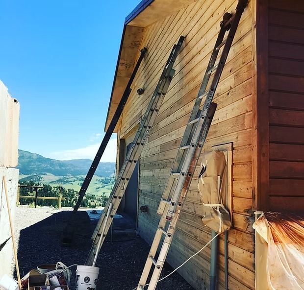 Two tall metal extension ladders lean against the unfinished wooden siding of a house under a clear blue sky.