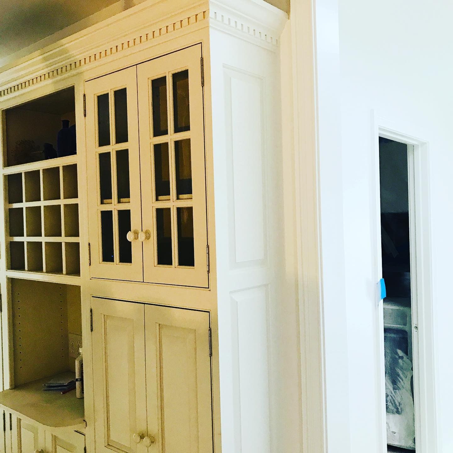 A tall, cream-colored wooden hutch with glass cabinet doors and a wine rack, standing next to a white doorway.