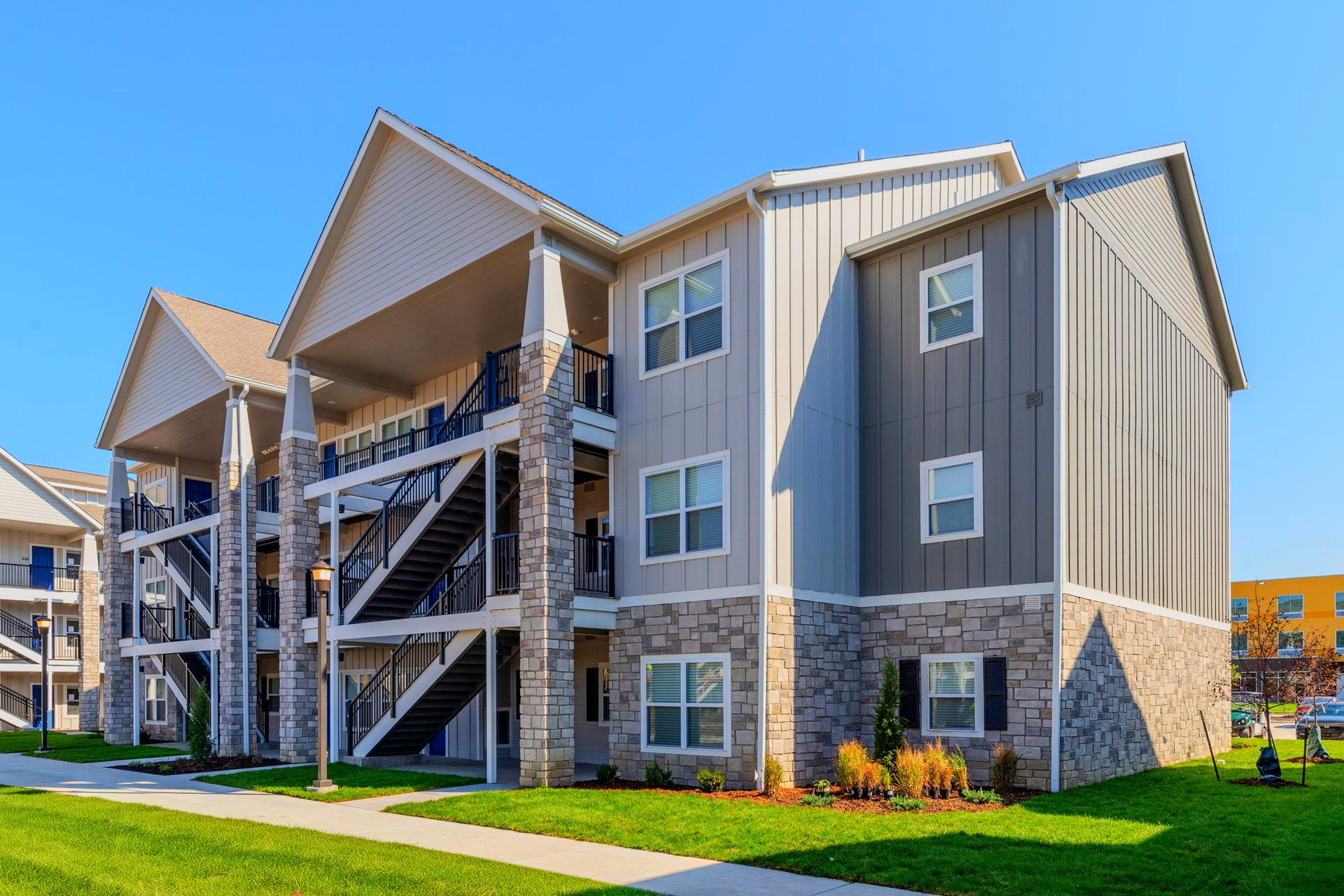 Three-story apartment building with stone facade, gray siding, and exterior staircases under a blue sky.