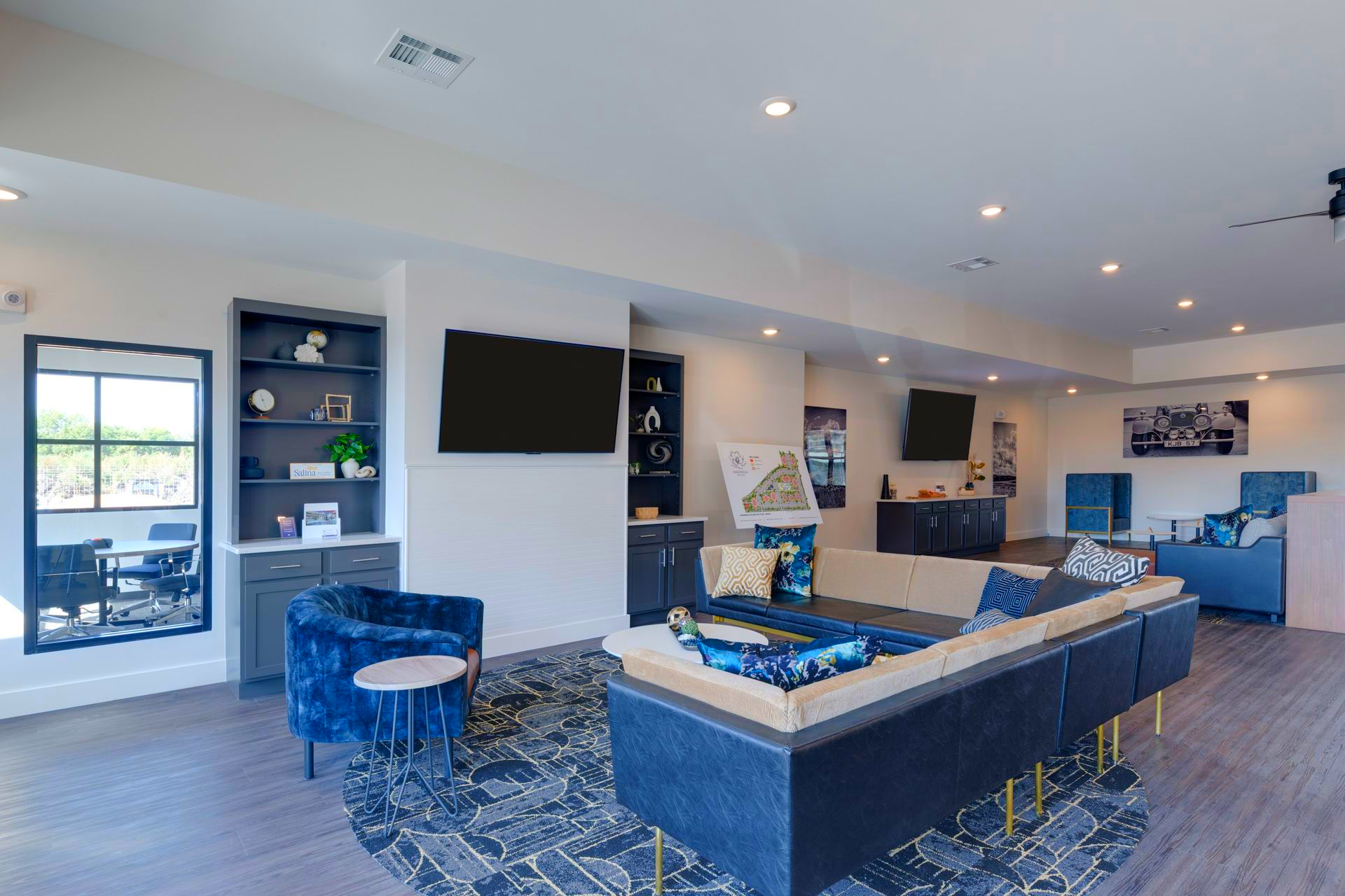 Spacious living area with blue sofas, armchairs, and a large rug; multiple TVs on the walls.