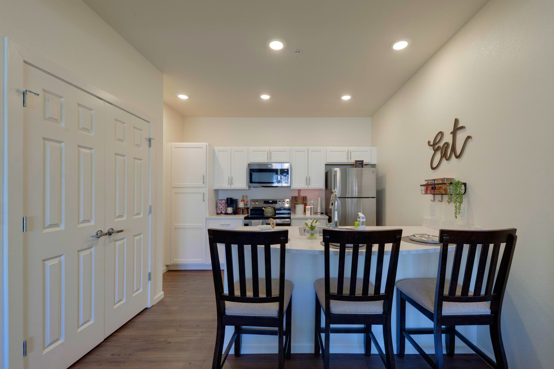 Kitchen with a white island and three black chairs. Cabinets, stainless steel appliances, and wood flooring.