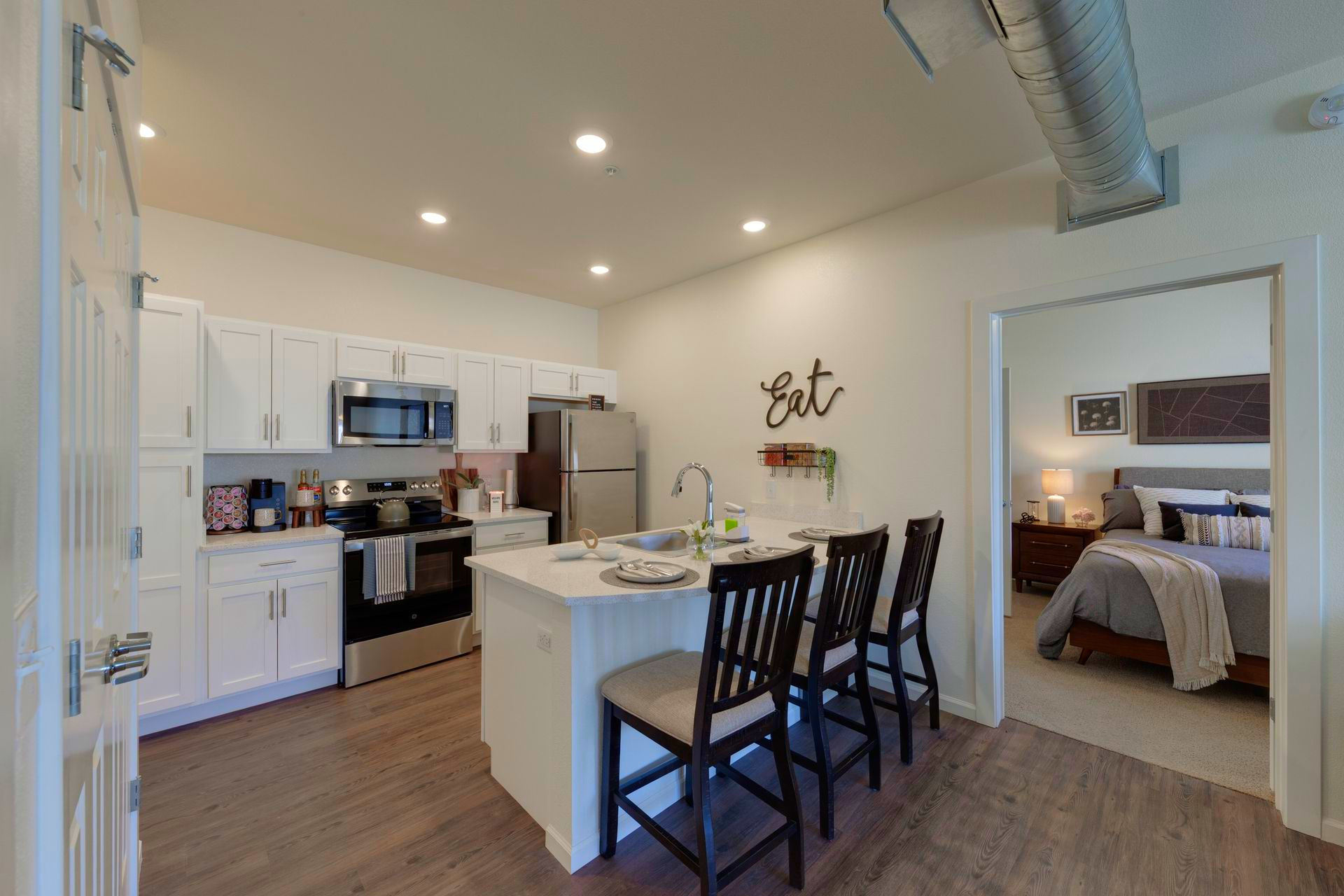 Modern kitchen with white cabinets, stainless steel appliances, island with seating, and a view into a bedroom.