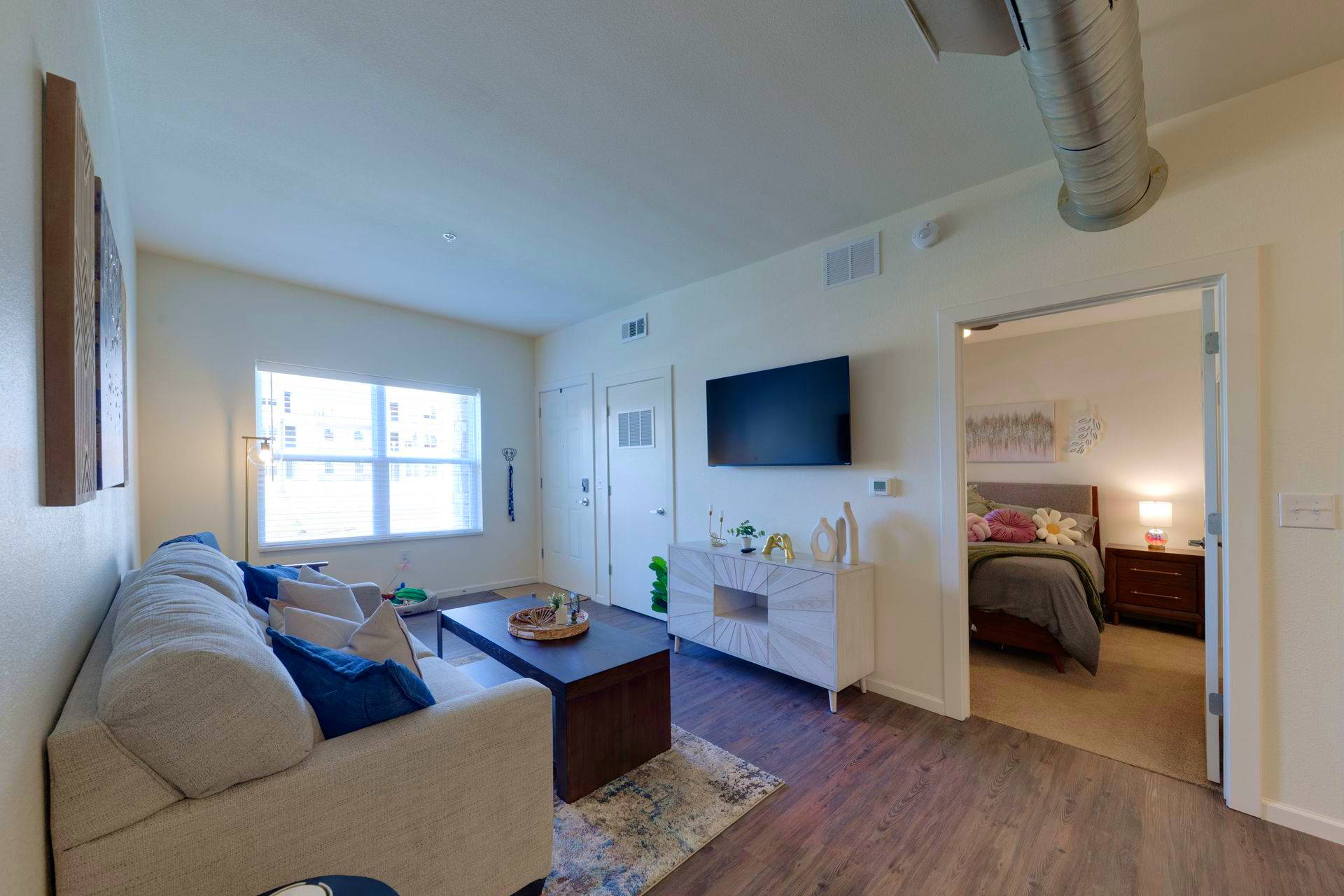 Cozy living room with sofa, TV, and bedroom doorway. Bright with neutral tones and wooden floors.
